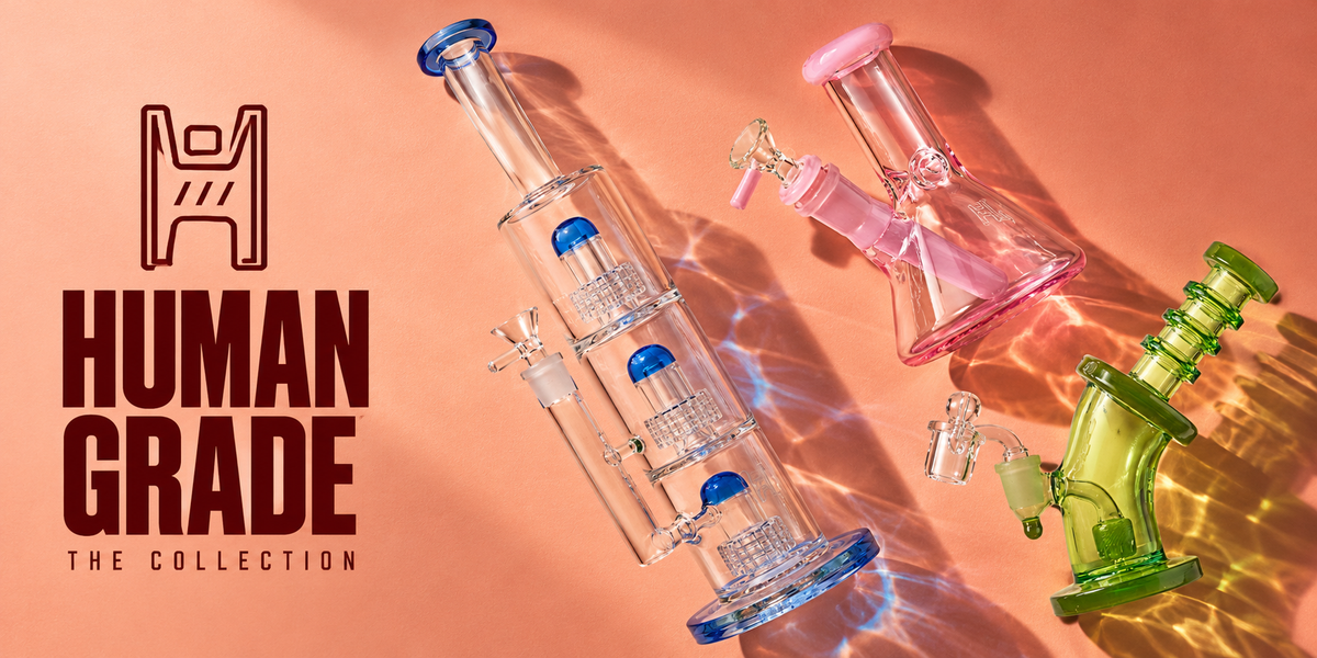 Human Grade glass in sunlight: a blue triple-perc tube, pink mini beaker and green bent-neck dab rig beside the logo