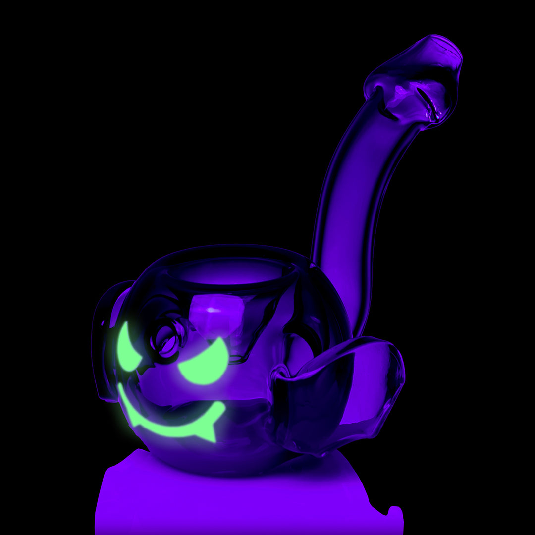 MJ Arsenal Bud-Sucker Hand Pipe under black light, the green face glowing, three-quarter view